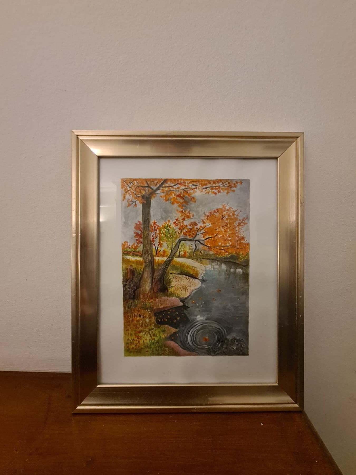 A gold-framed watercolour of trees in autumn colour along a still river, the far bank reflected in the water.