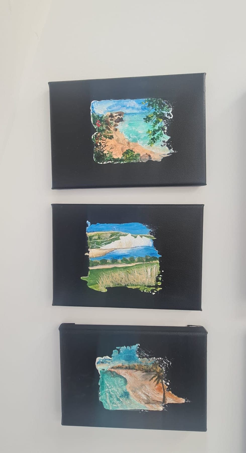 Three small textured coastal studies hung in a vertical column on a white wall, each mounted on a dark board — sea, sand and headland in each one.