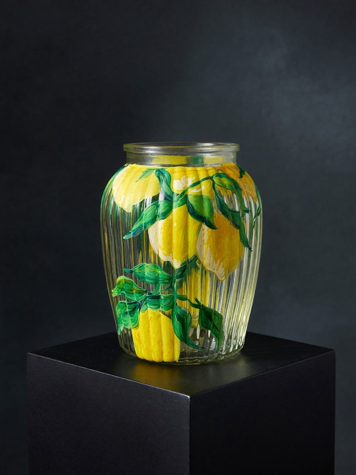 A ribbed clear glass vase hand-painted with bright yellow lemons and deep green leaves, lit against a black background.