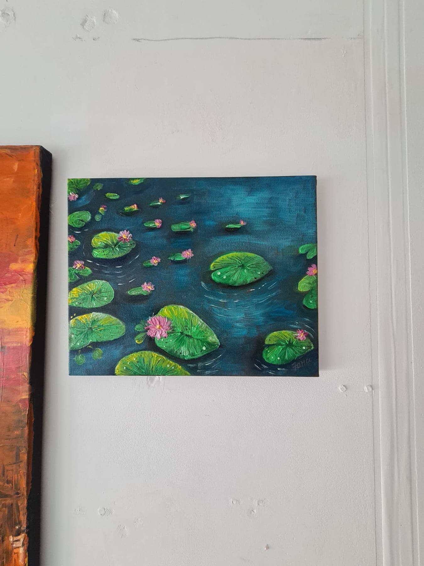 A canvas hung on a white wall showing water lilies and flat round pads floating on deep teal-green water.