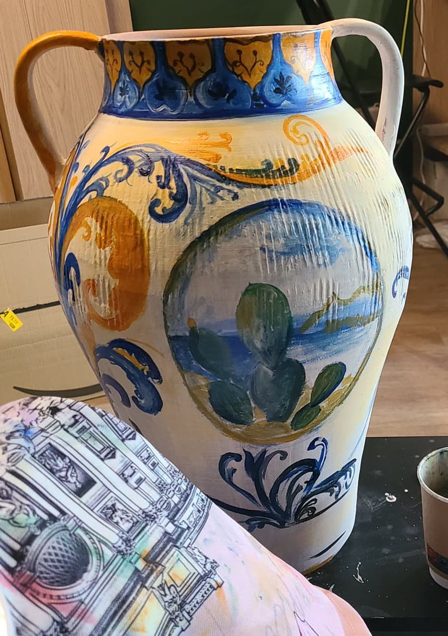 A tall two-handled earthenware urn hand-painted in the maiolica manner — blue and ochre scrollwork around a landscape cartouche of prickly pears above a bay.