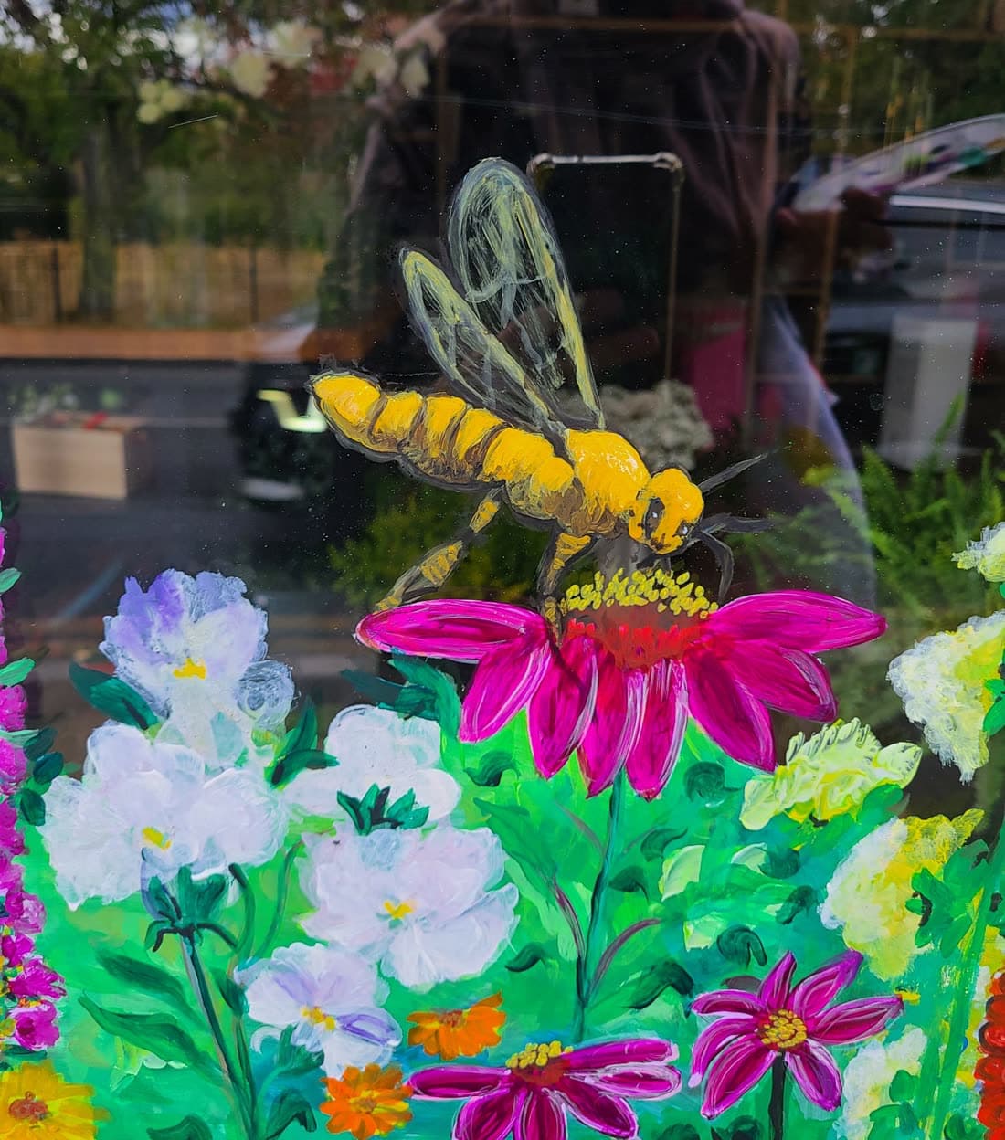A large bee painted on a shop window, wings left as transparent outlines, settling on a magenta echinacea above a bank of white and lilac blooms.