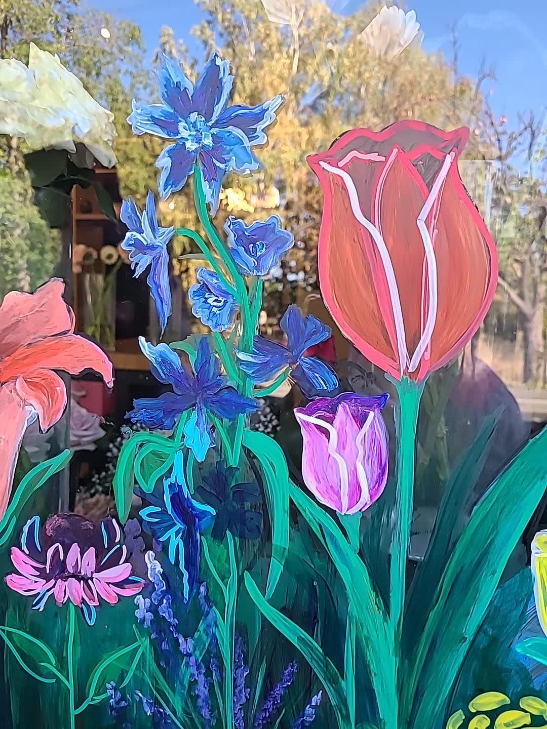 Blue delphiniums and a scarlet tulip painted on shop glazing, with the florist's shelves and the street showing through the clear glass behind.