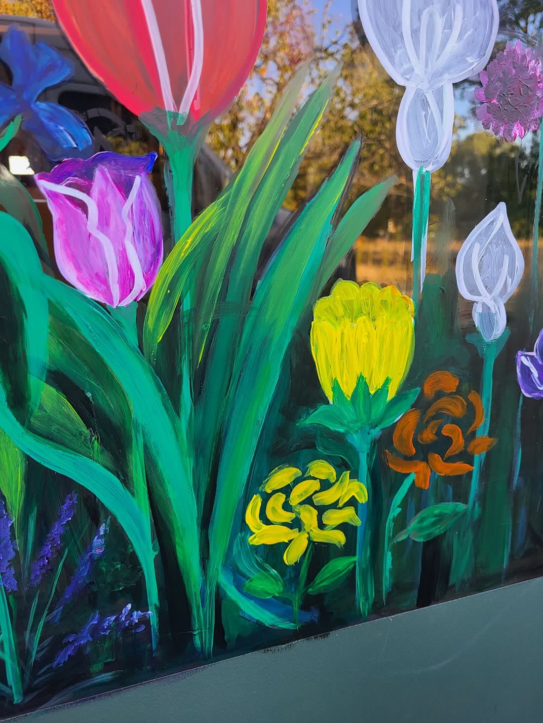 Pink and scarlet tulips, a yellow bloom and an orange rose painted on shop glazing, the leaves running down to where the paint stops at the sill.