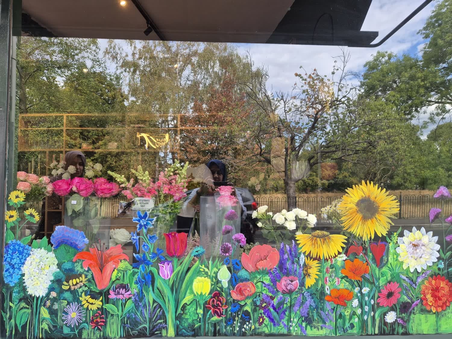 A florist's shop window painted end to end with a wildflower border — sunflowers, dahlias, tulips and delphiniums — with the real bouquets and the street both visible through the clear glass above.