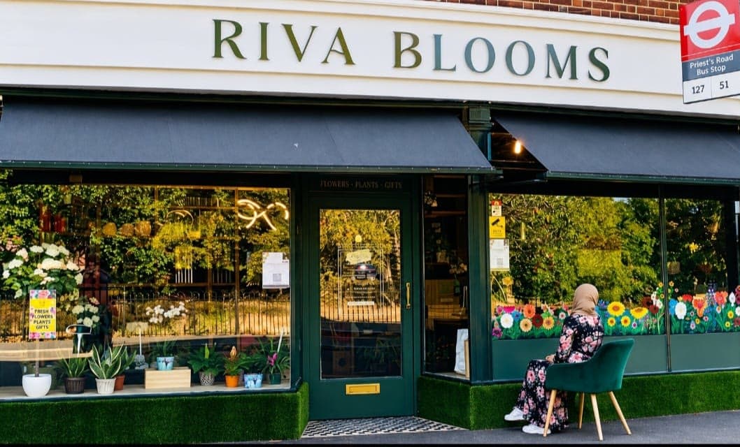 A florist's shopfront with a hand-painted wildflower border running along the bottom of its window glazing, and the artist sitting outside on a green velvet chair.