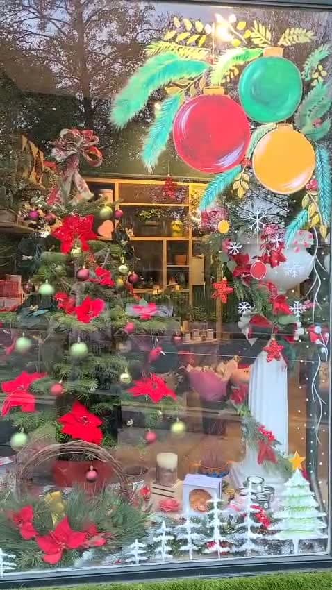 Painted baubles and a teal pine spray on a florist's window at dusk, with poinsettias, a lit Christmas tree and a white fluted plinth behind the glass.